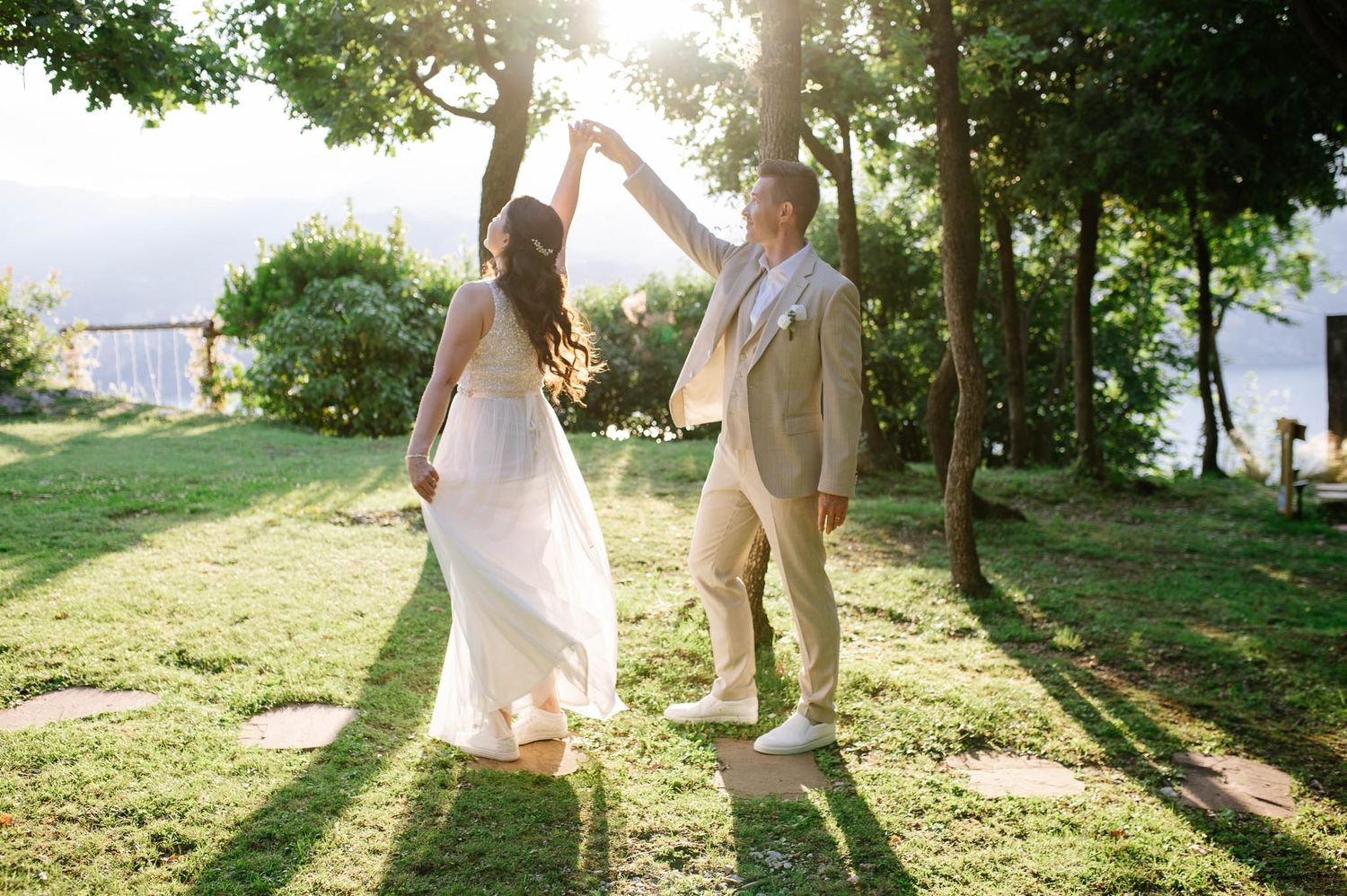Bride and groom dancing joyfully on green lawn surrounded by trees with golden sunlight glowing.