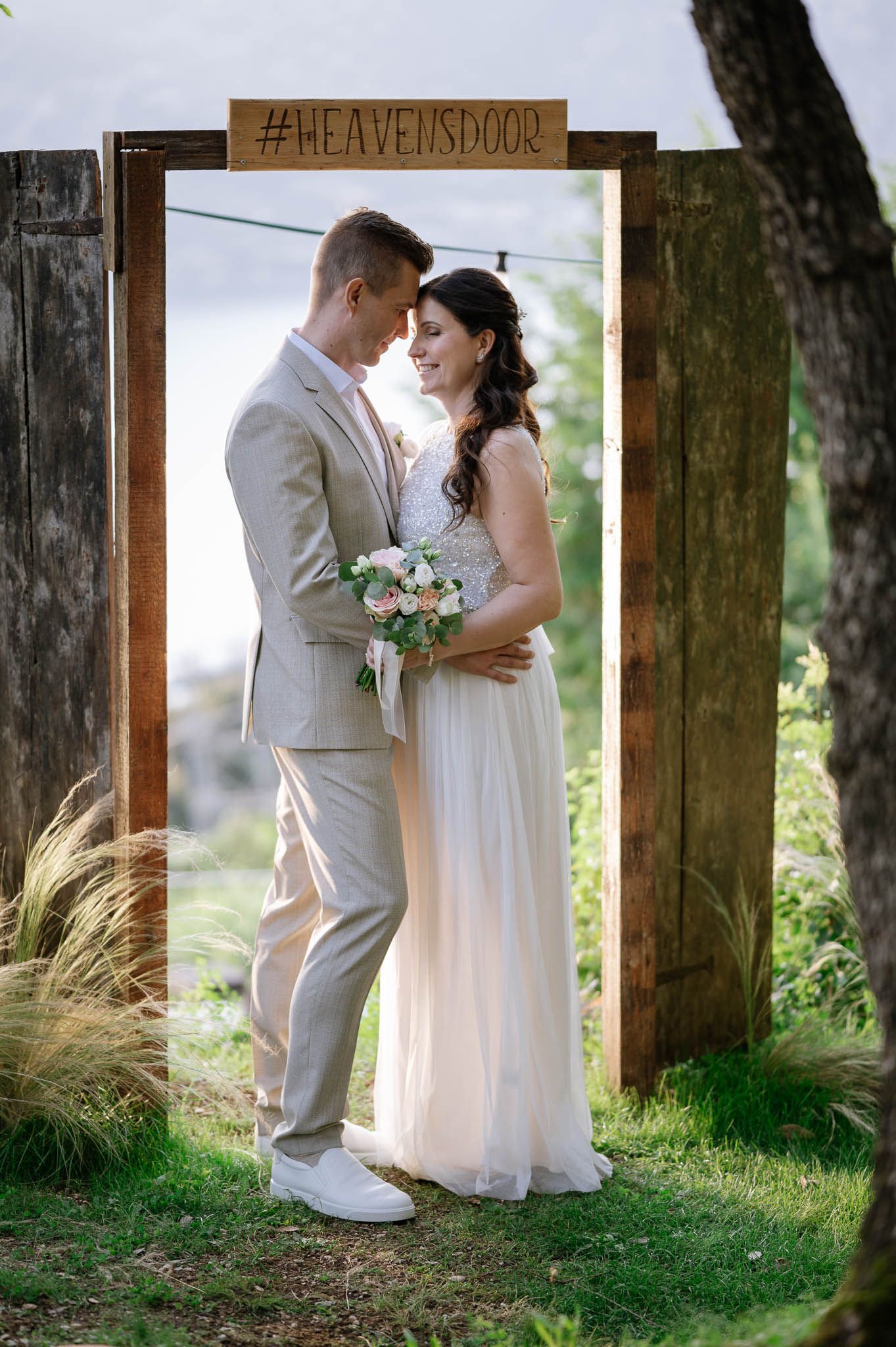 Bride and groom embrace under rustic wooden arch with #HEAVENSDOOR sign, bride holding pink bouquet in white gown.