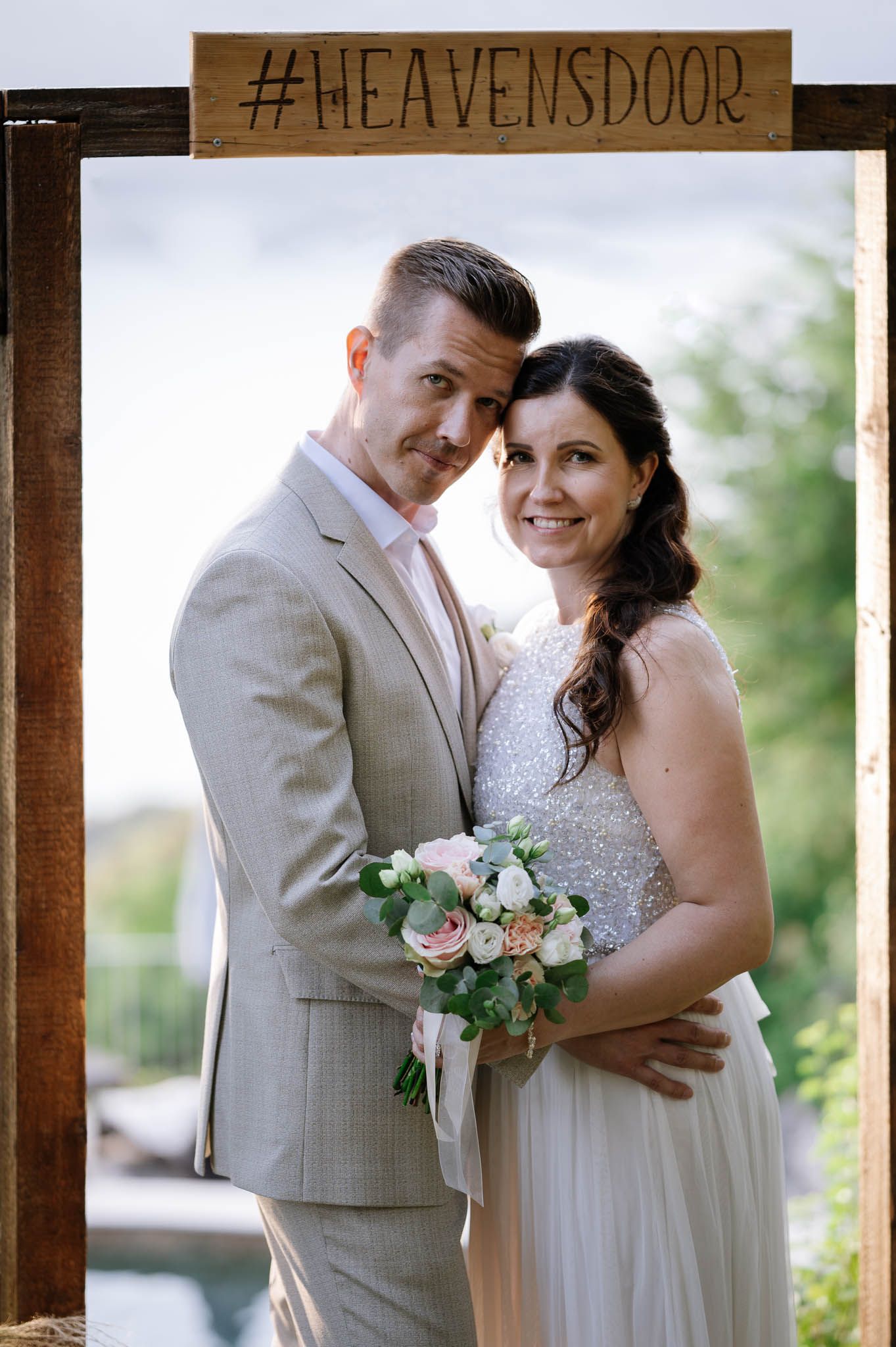 Bride and groom posing under a wooden arch with #HEAVENSDOOR sign, bride holding pink bouquet at outdoor wedding.