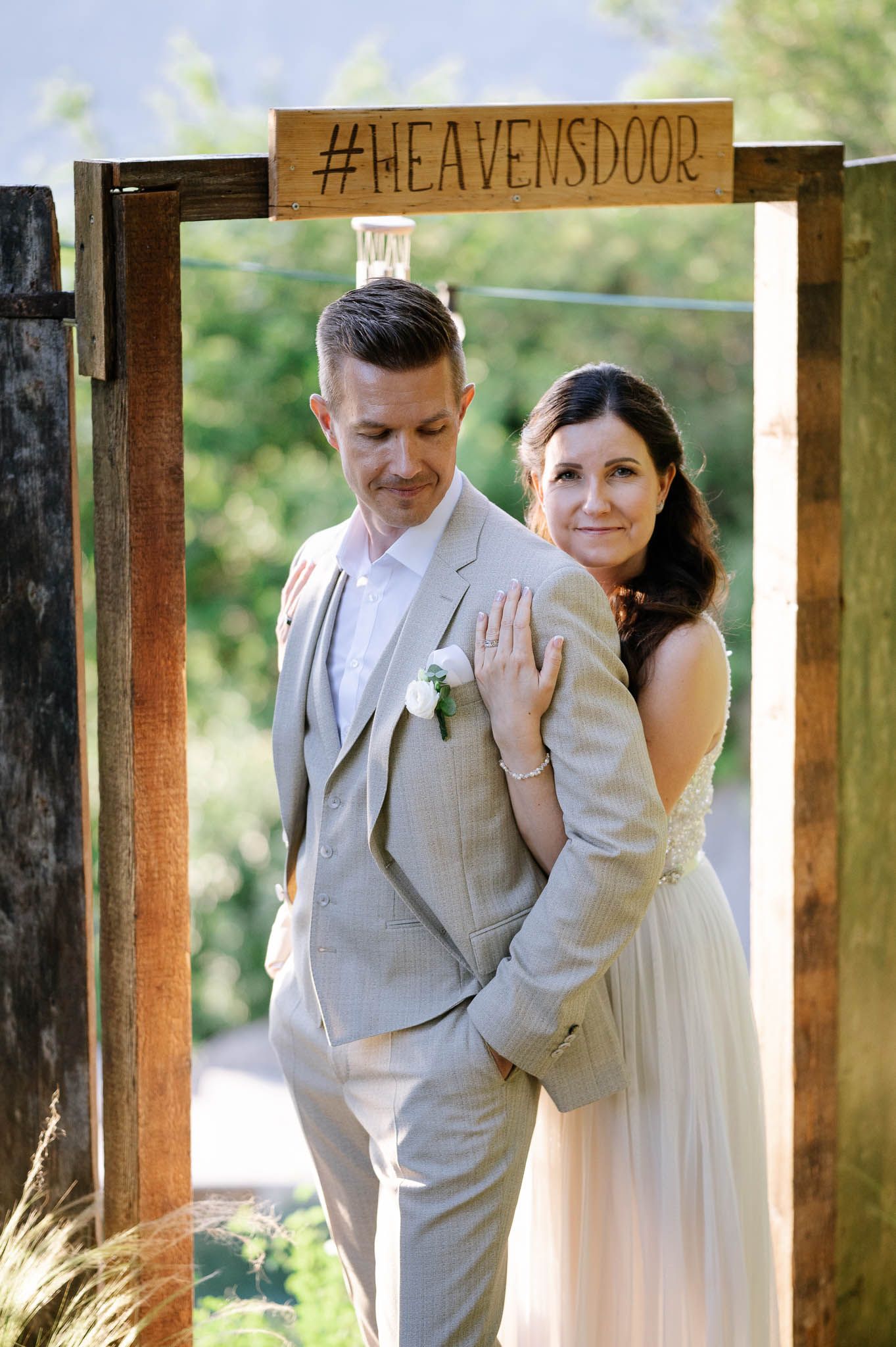 Bride and groom posing under a rustic wooden arch with #HEAVENSDOOR sign at an outdoor wedding.