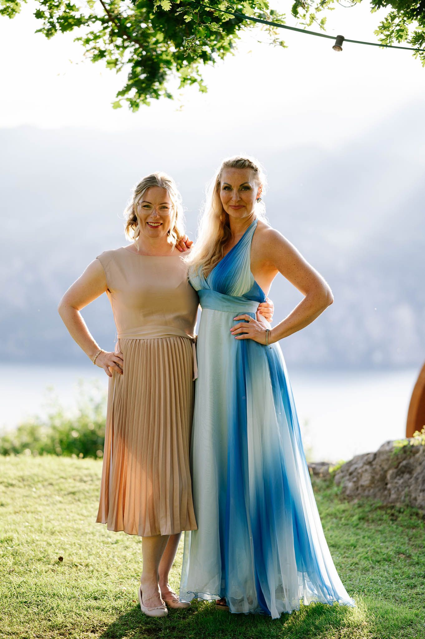 Two women in elegant dresses pose outdoors near a lake, one in beige pleated skirt, other in blue ombre gown.