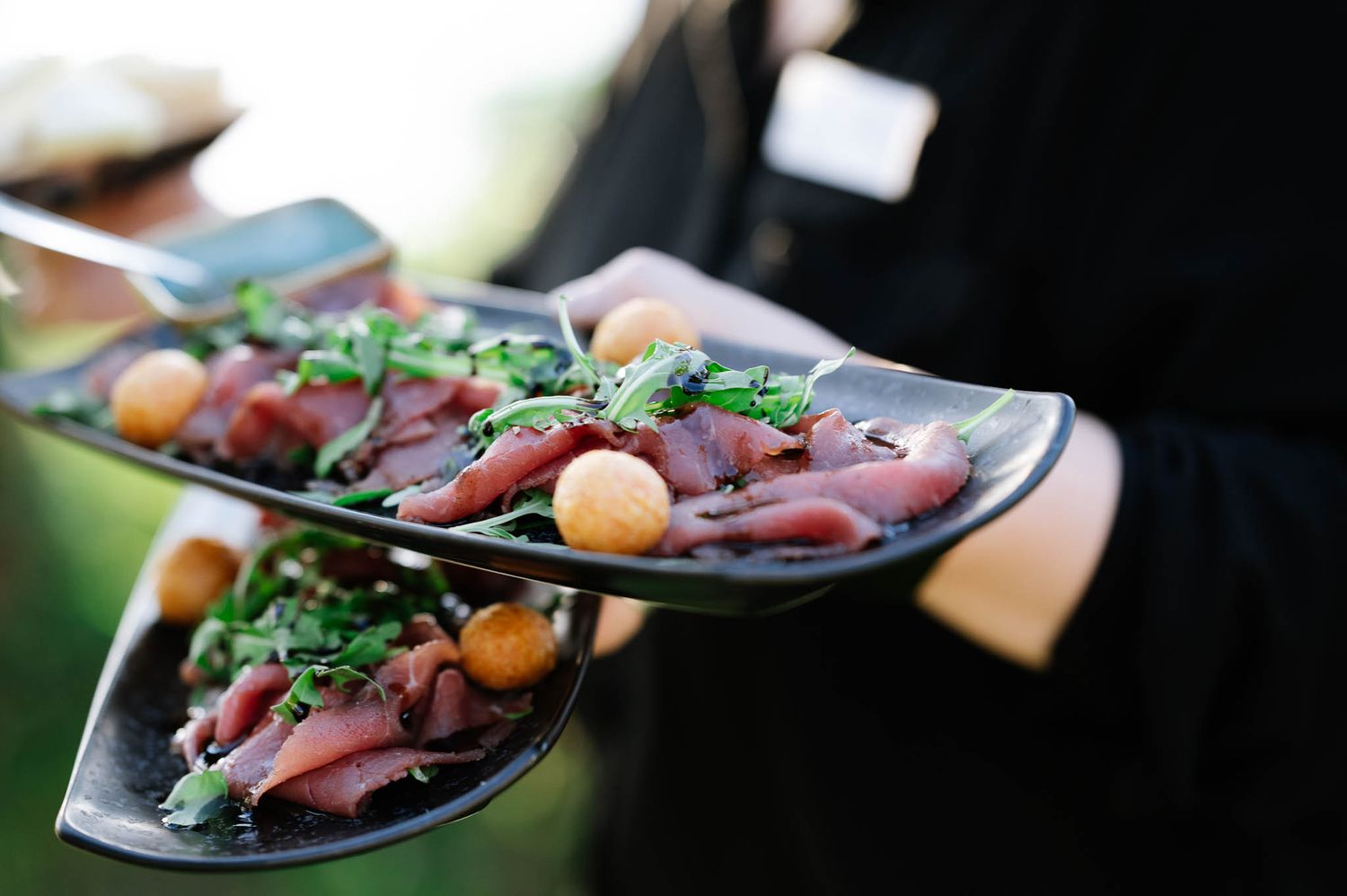 Catering server holding two elegant black plates with gourmet appetizers garnished with fresh herbs and crispy bites.