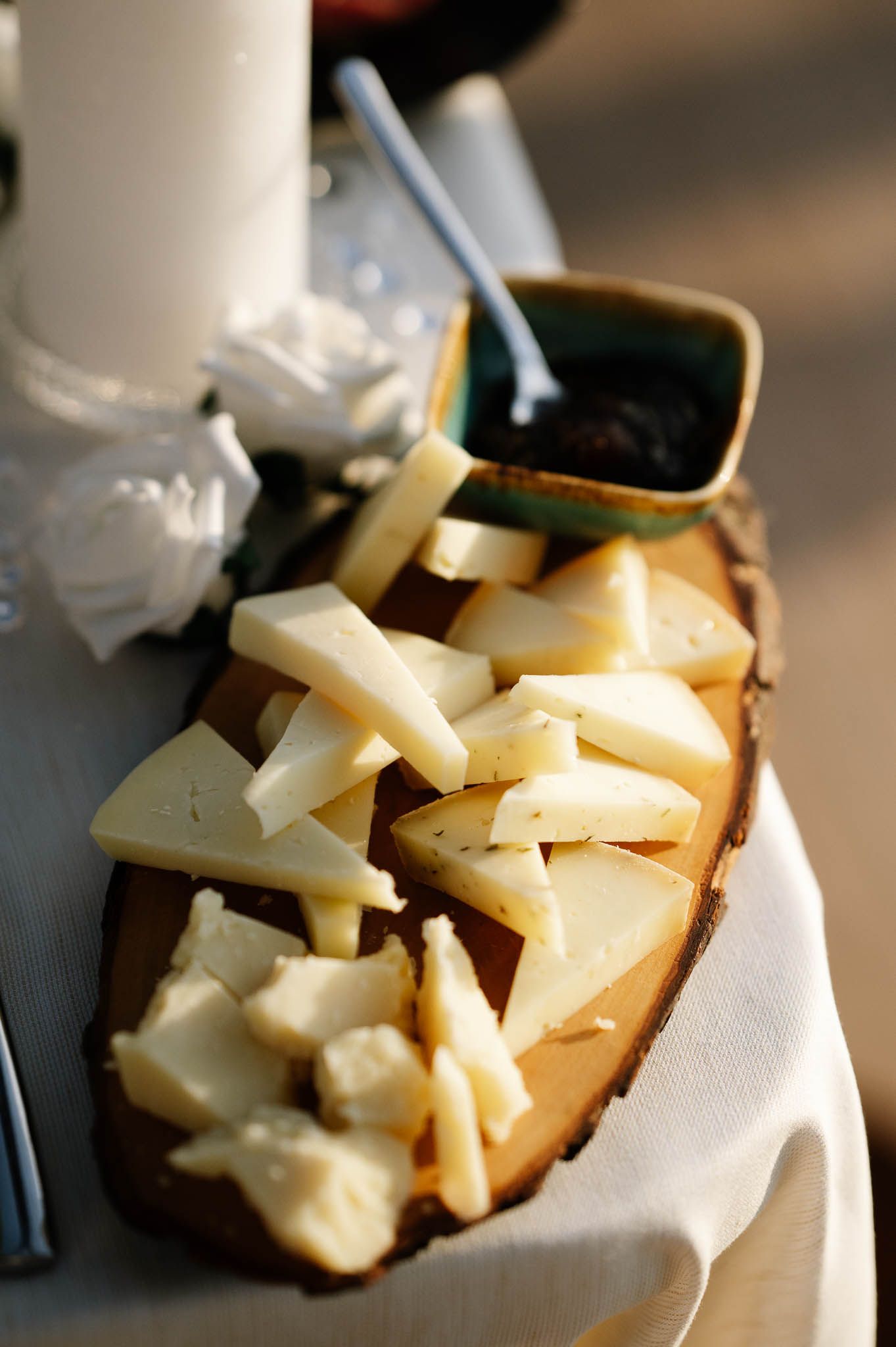 Assorted cheese slices arranged on a rustic wooden board with a small dish of accompaniments on a white tablecloth.