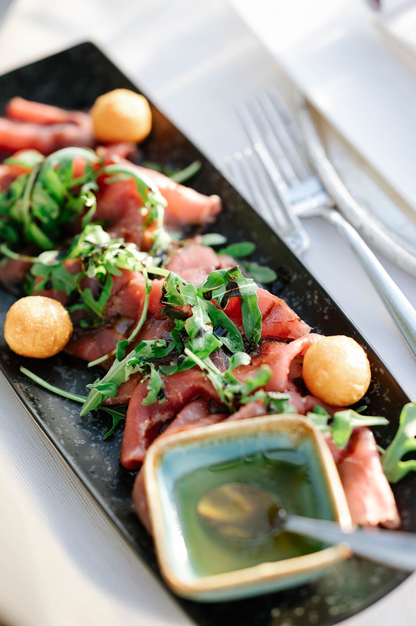 Elegant appetizer plate with cured salmon, arugula, melon balls, and herb dipping sauce on a black rectangular dish.