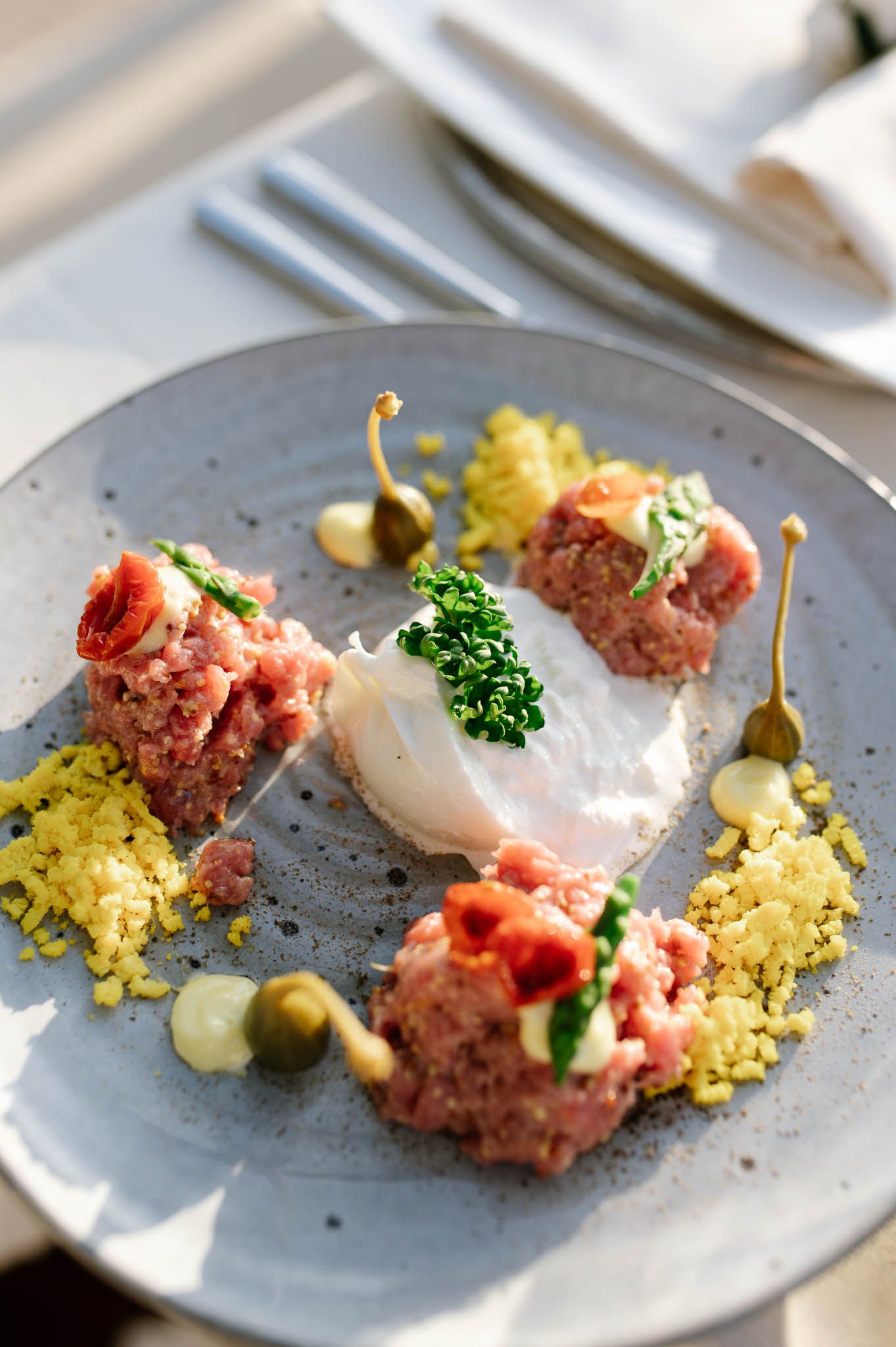 Elegant beef tartare with capers, herbs, and egg on a gray plate, fine dining presentation.