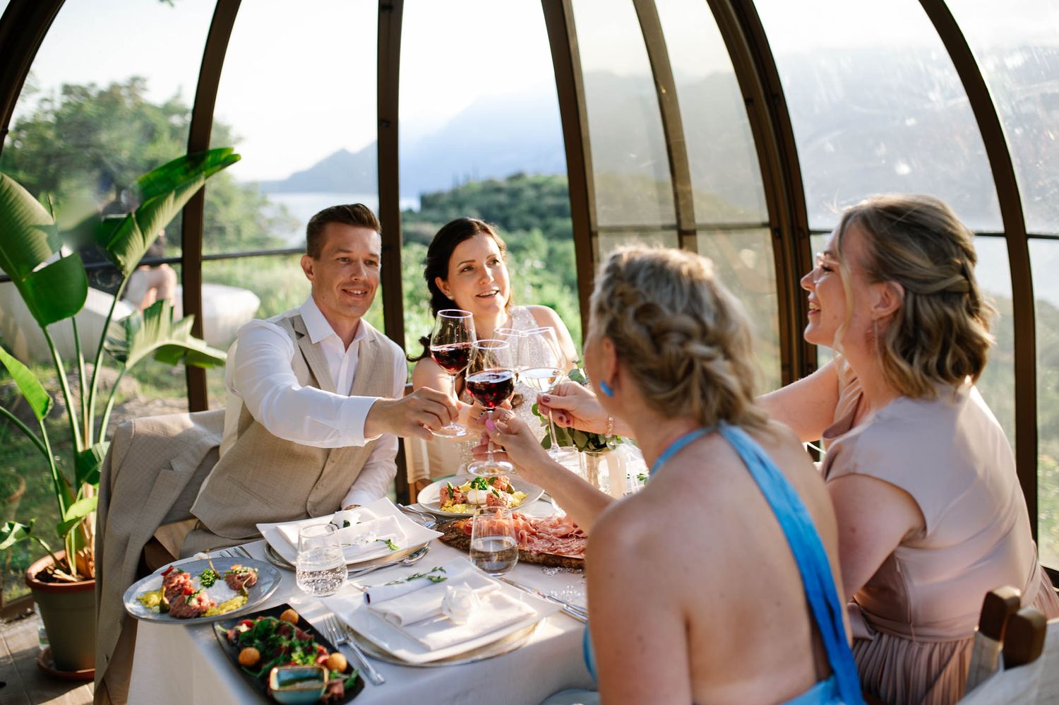 Four guests toasting with red wine at an elegant glass-enclosed restaurant with scenic mountain views.