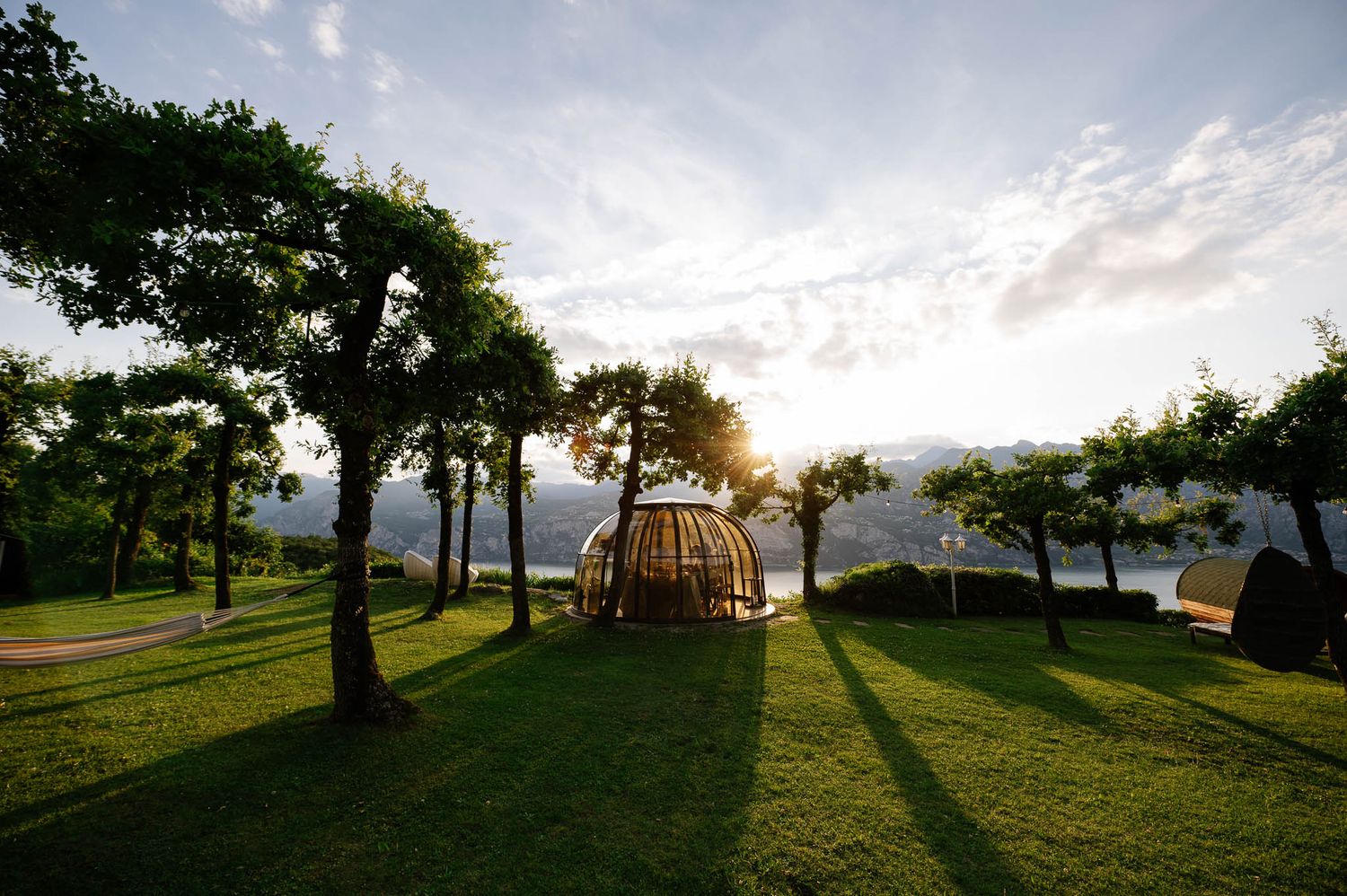 Wooden dome structure on lush green lawn surrounded by trees with sunlight streaming through at golden hour near a lake.