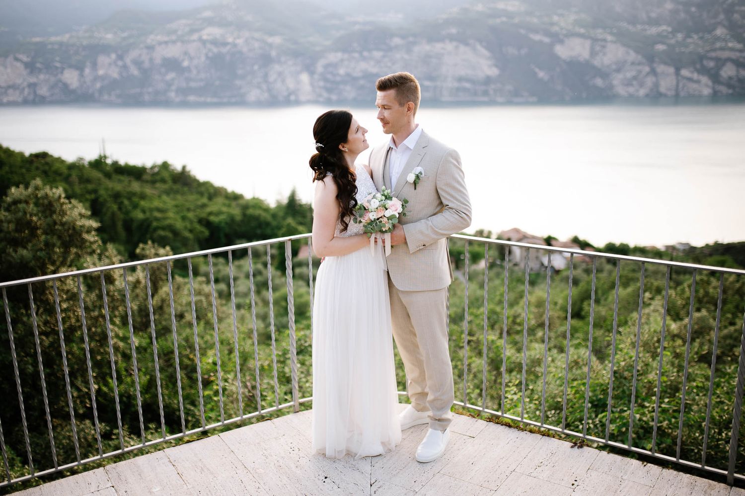 Bride and groom share a romantic moment on a terrace overlooking lush hills and a lake on their wedding day.