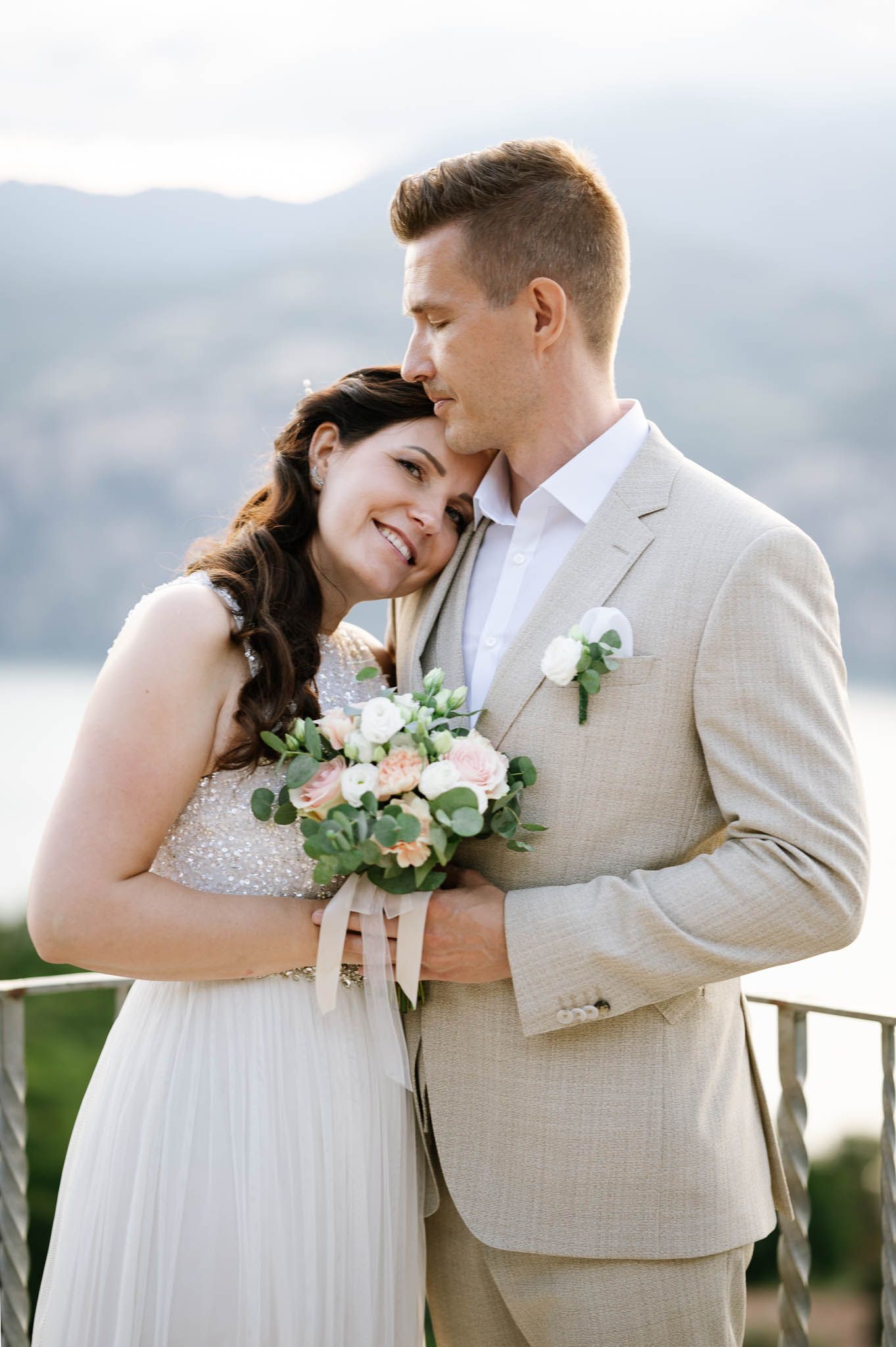 A bride and groom share a tender moment outdoors, she holds a blush bouquet, mountains visible behind them.