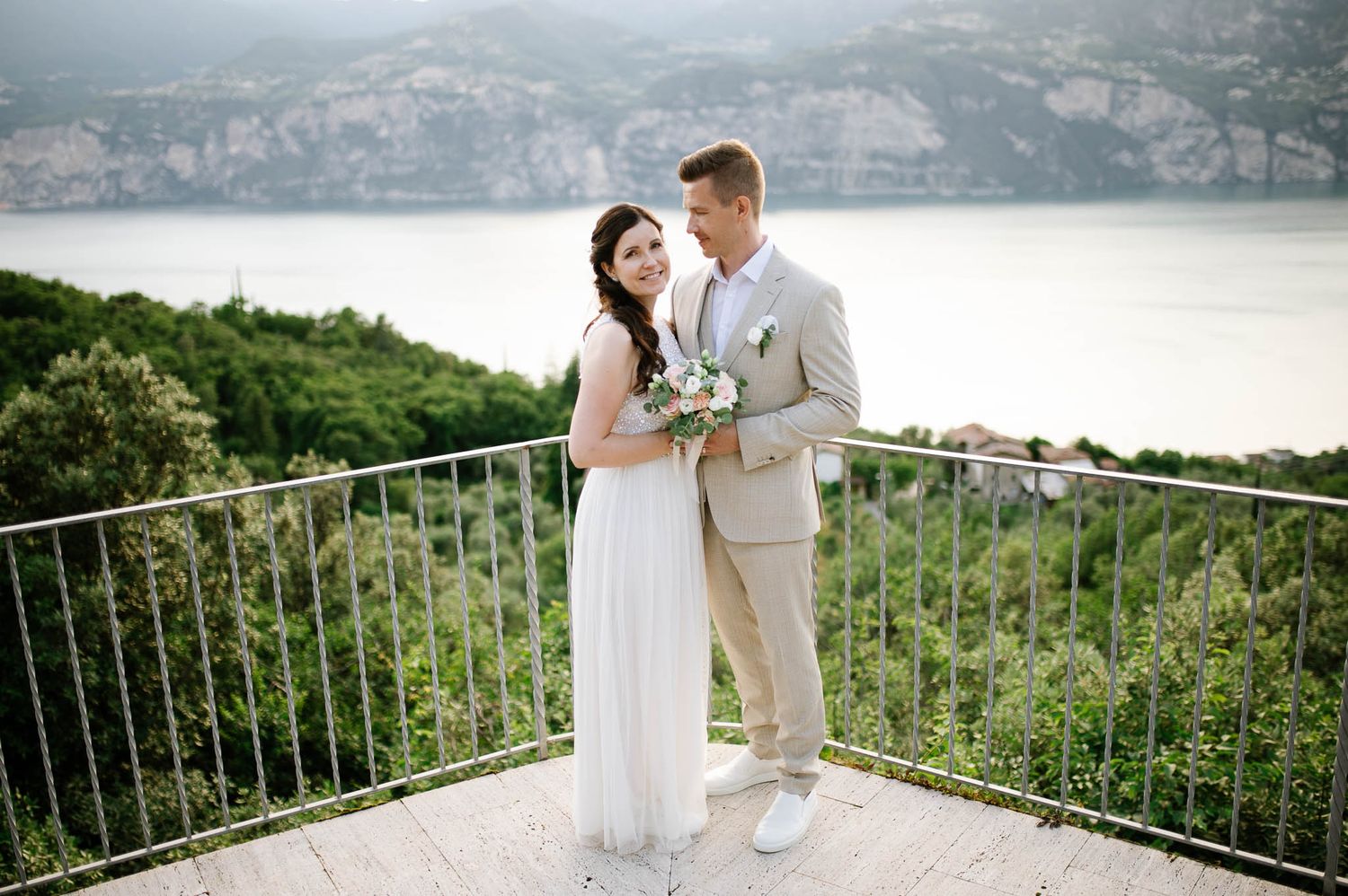 Bride and groom pose on a scenic balcony overlooking Lake Garda, surrounded by lush greenery on their wedding day.