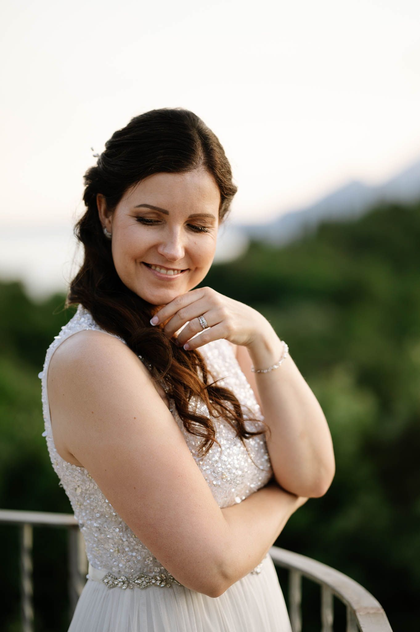 Smiling bride in beaded white gown with diamond ring, standing on balcony with lush greenery at golden hour.