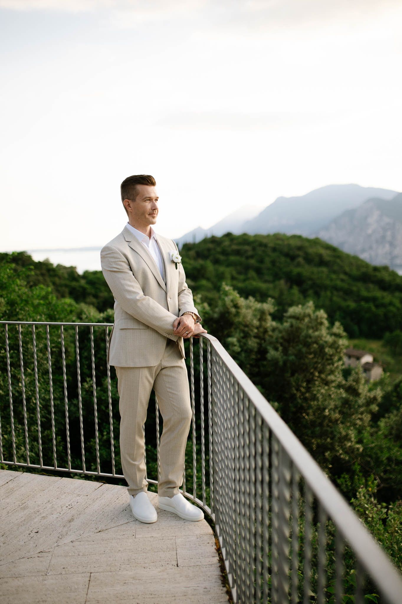 Groom in beige suit standing on scenic balcony overlooking lush green mountains and lake at dusk.
