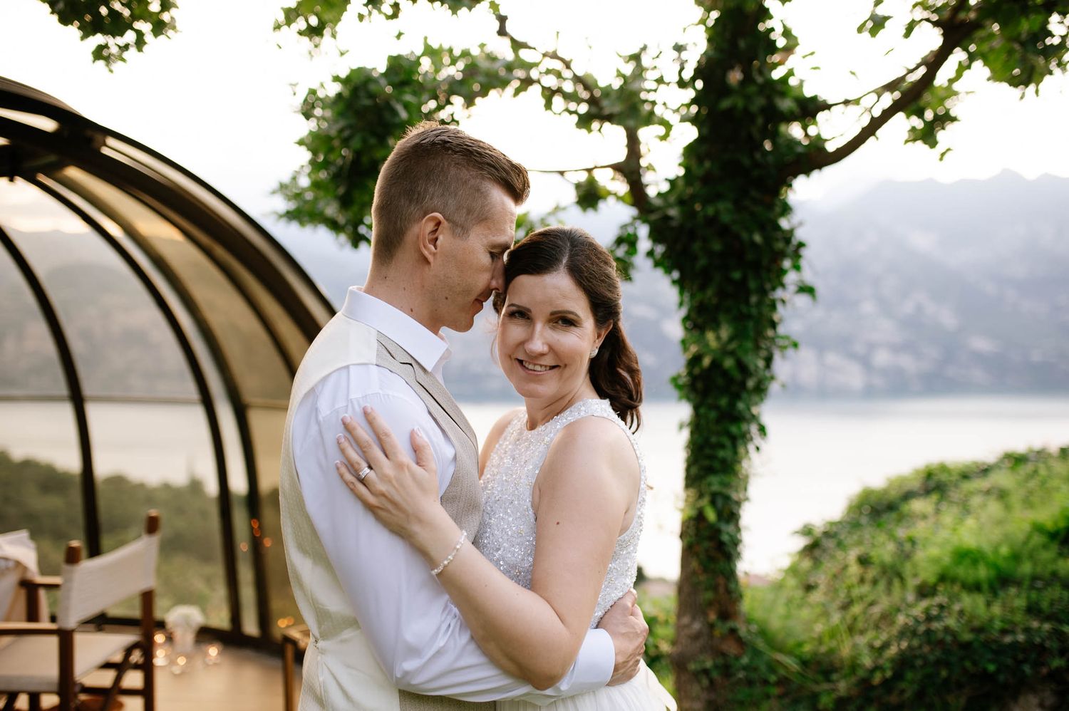 Bride and groom embracing outdoors near a lake with lush greenery and mountains in the background.