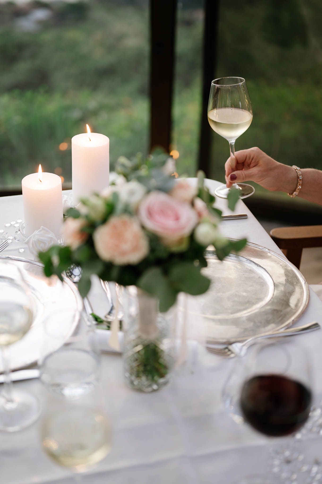 Elegant wedding table setting with white candles, pink roses, silver charger plates, and a hand holding a glass of white wine.