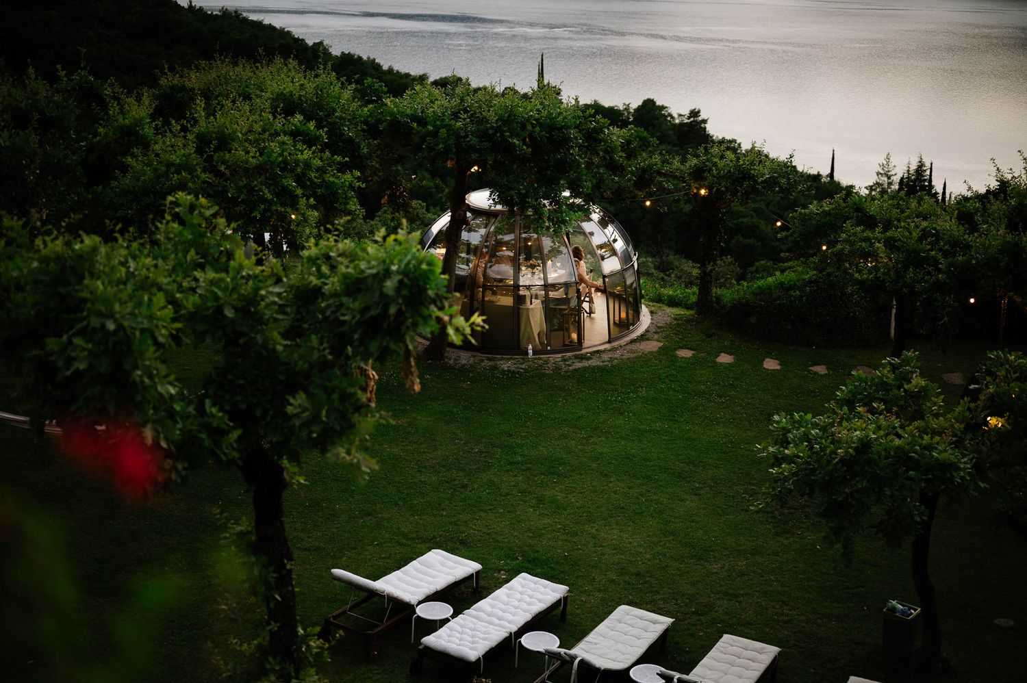 Aerial view of illuminated geodesic dome greenhouse in lush garden with white chairs at twilight.