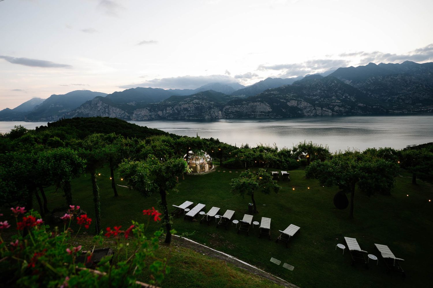 Stunning aerial view of elegant lakeside wedding venue with white chairs and mountain backdrop at dusk.