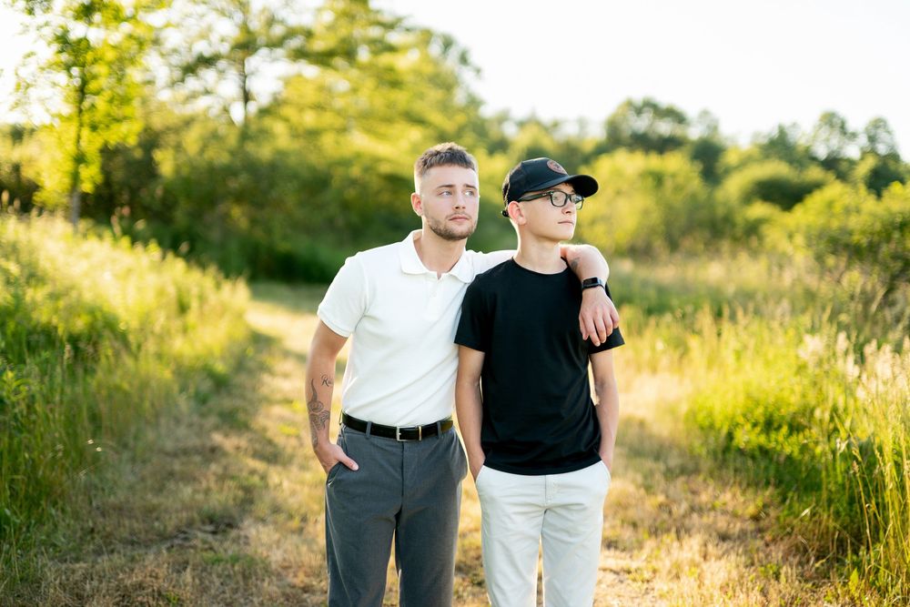 Two young men pose together on a sunny nature path, one in white polo and gray pants, other in black tee and cap.