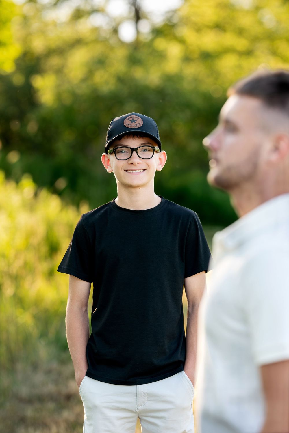 Smiling boy in black t-shirt, cap, and glasses stands outdoors with blurred man in foreground.