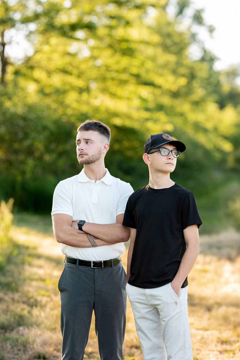 Two young men standing outdoors in summer, one in white polo with crossed arms, other in black tee and cap.