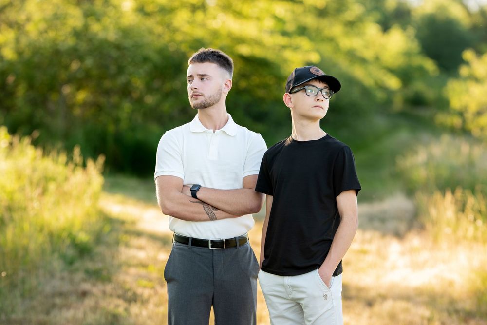 Two young men standing outdoors on a sunny day, one in white polo with crossed arms, other in black tee and cap.