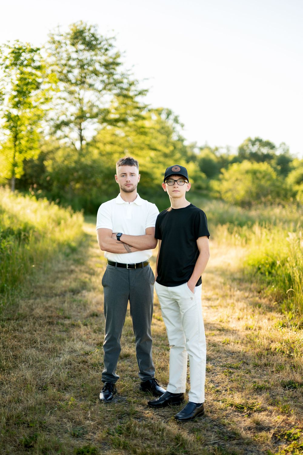 Two young men pose formally in a sunny field, one in white polo with crossed arms, other in black tee and baseball cap.