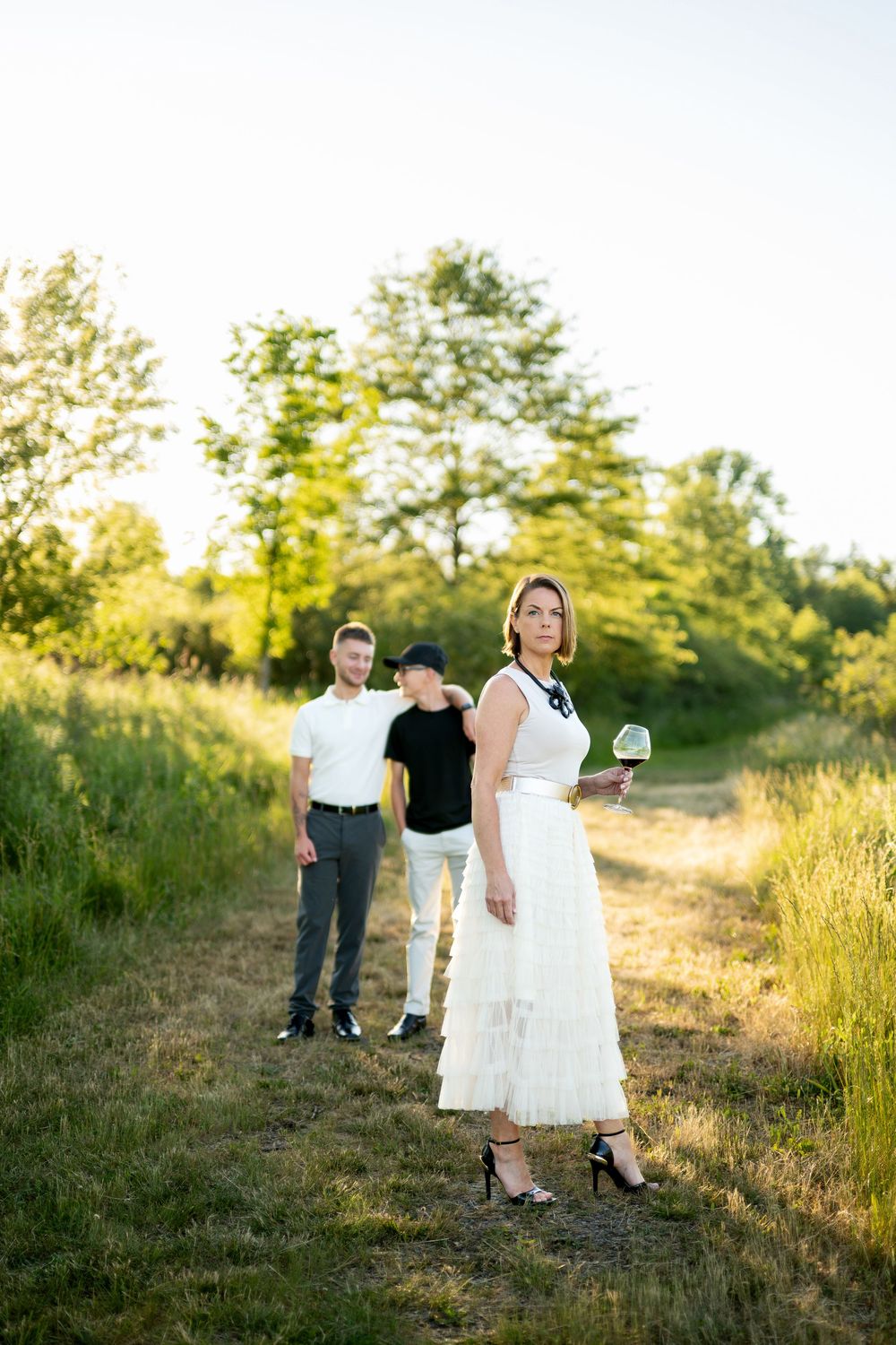 Woman in white dress holding a flower in a sunny green field with two men standing behind her.