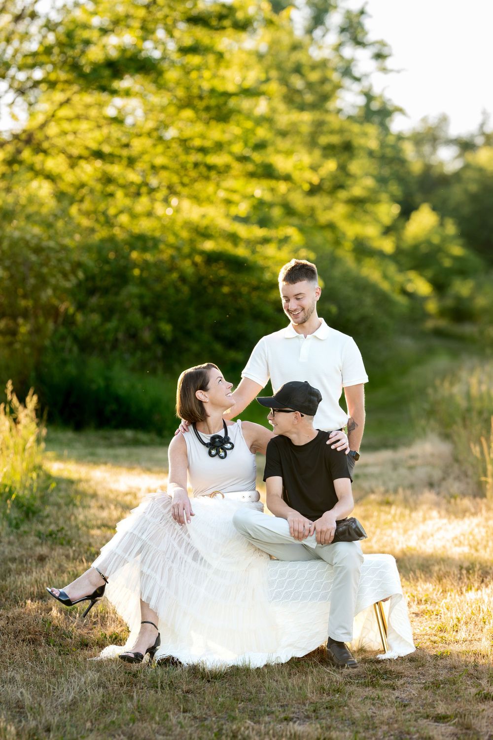 Family of three enjoying sunny outdoor photo session, mother in white dress seated with son and teenage boy standing behind.