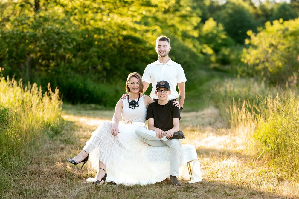 Happy family of three posing in sunny meadow, mother in white dress seated with two sons standing nearby.