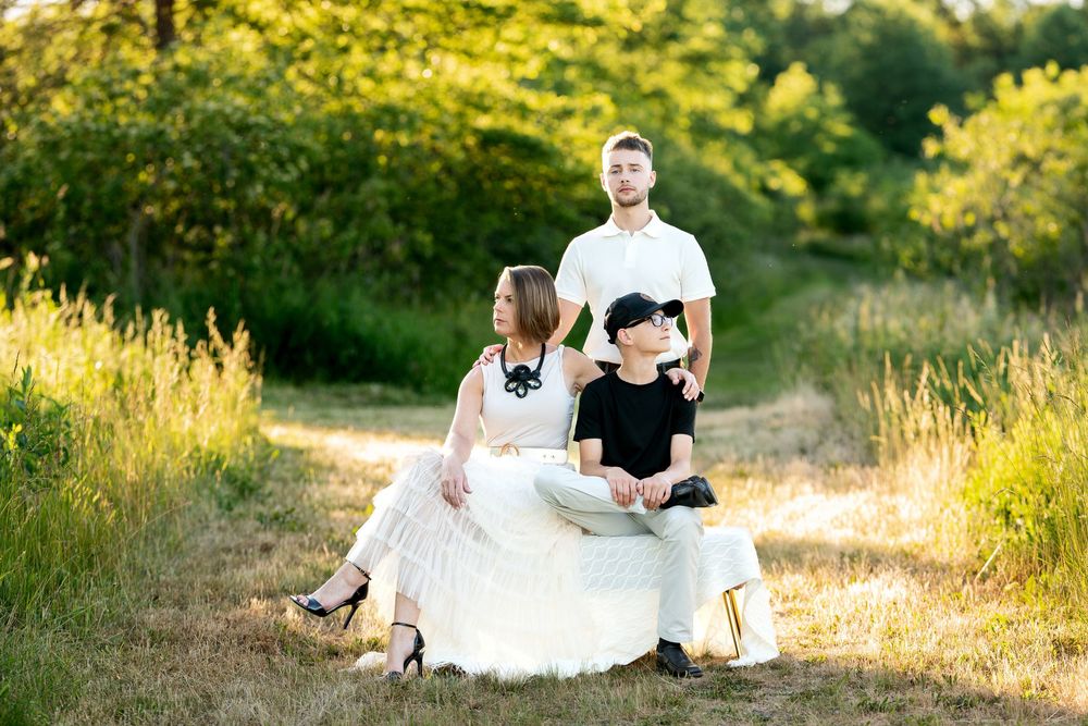 Family of three sitting together on a sunny meadow path surrounded by lush green trees.