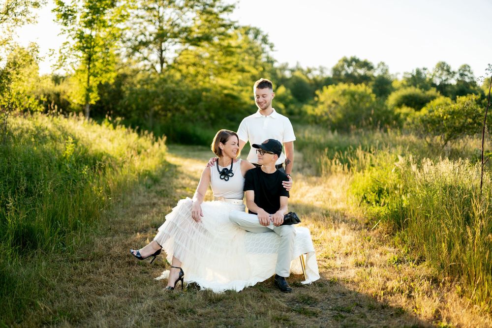 Happy family of three posing together on sunny dirt path surrounded by lush green meadow and trees.