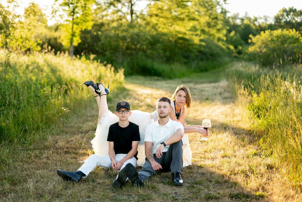 Three people sit relaxed on grass in golden sunlight, one in white dress, others in casual clothes, holding wine glass.