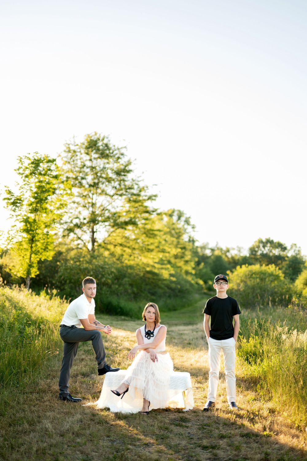 Three people posing outdoors in a sunny field, woman in white dress seated, two men standing beside her.