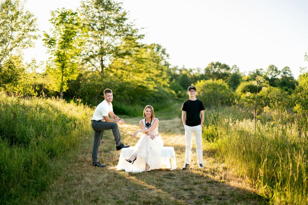 Playful family photo outdoors in golden sunlight, two men and a woman in white dress laughing on a green field path.