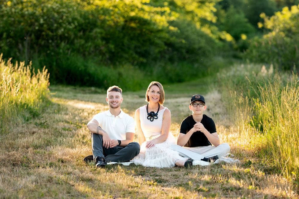 Happy family of three sitting on grass in a sunlit field with green trees in background.