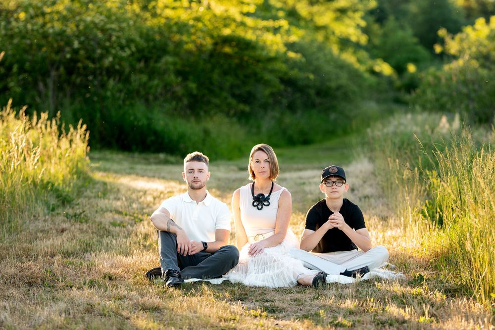 Three people sit cross-legged on grass in a sunlit field with green trees behind them.