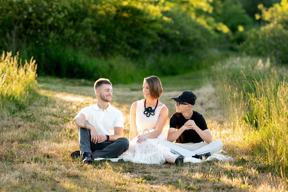 Family of three sitting together on dry grass in a sunny field, sharing a relaxed and candid outdoor moment.