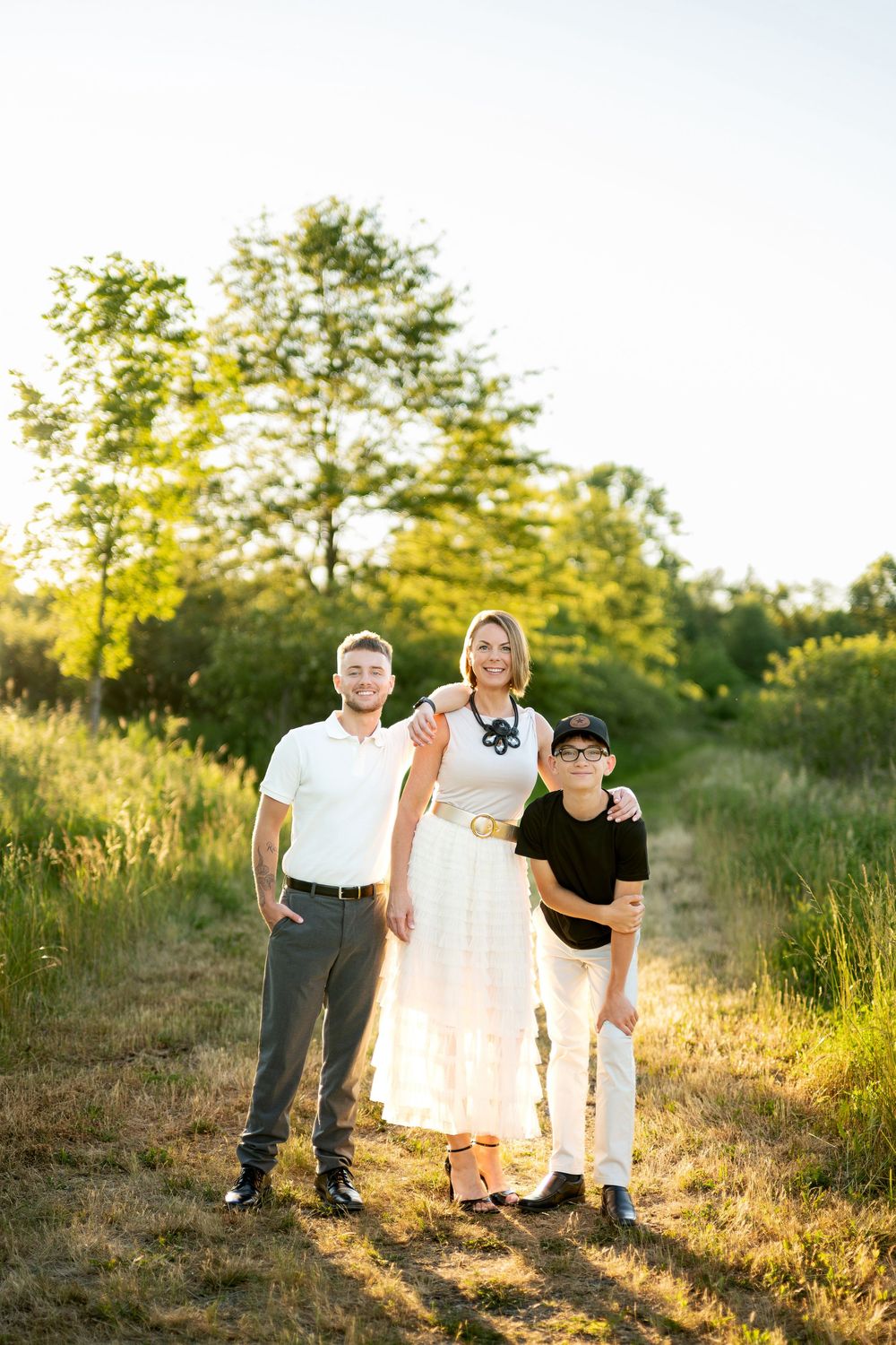 Happy family of three posing on a sunny nature path, mother in white dress flanked by husband and son.