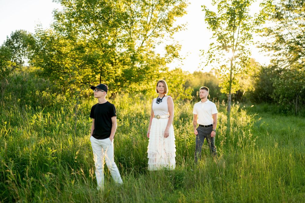 Family of three standing in lush green meadow during golden hour, mother in white dress with two sons.