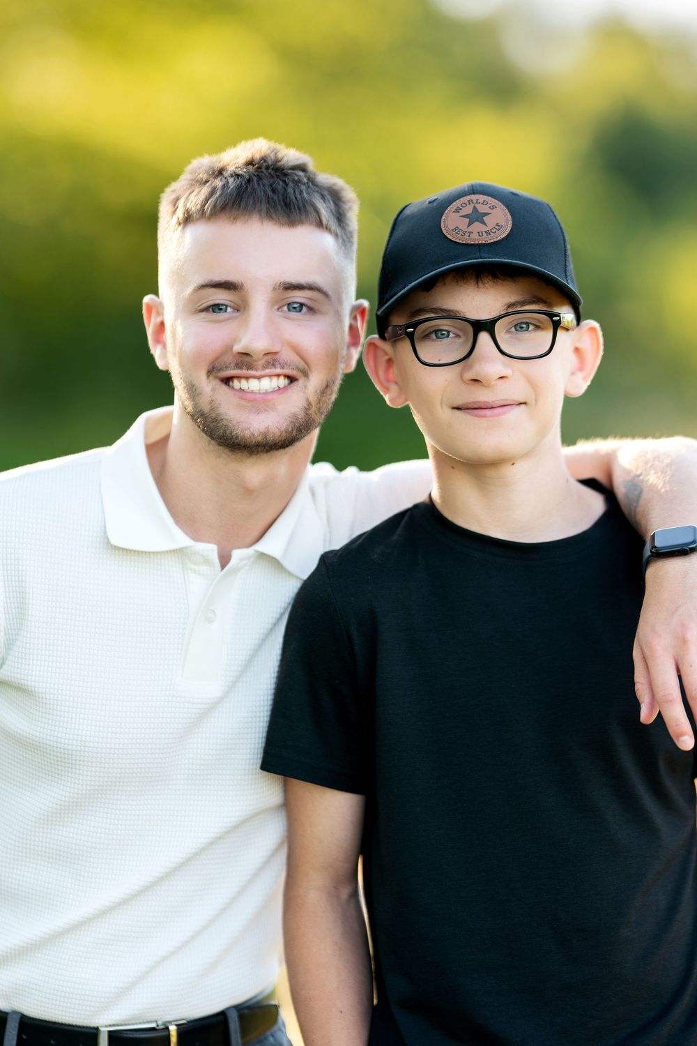 Two smiling young men pose together outdoors in warm sunlight, one in white polo, other in black tee and Converse cap.