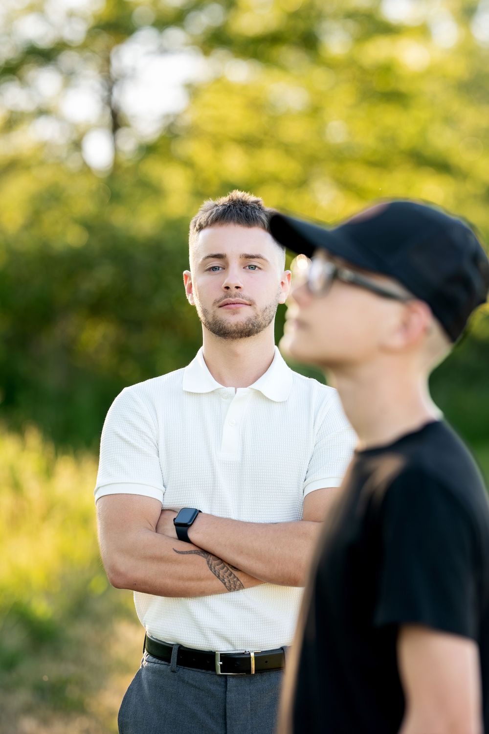Young man in white polo shirt standing with arms crossed outdoors in nature.