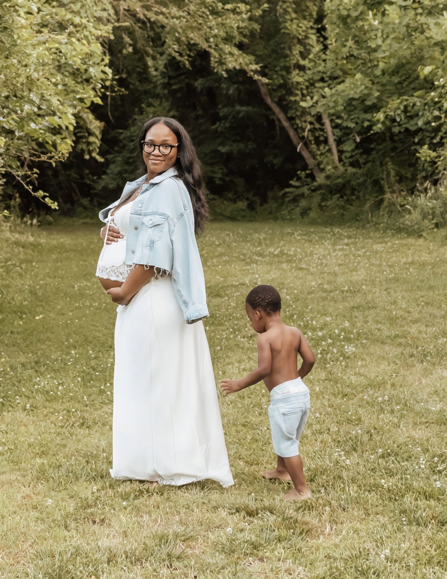Pregnant woman in white dress and denim jacket poses outdoors with young toddler boy on green grass lawn.