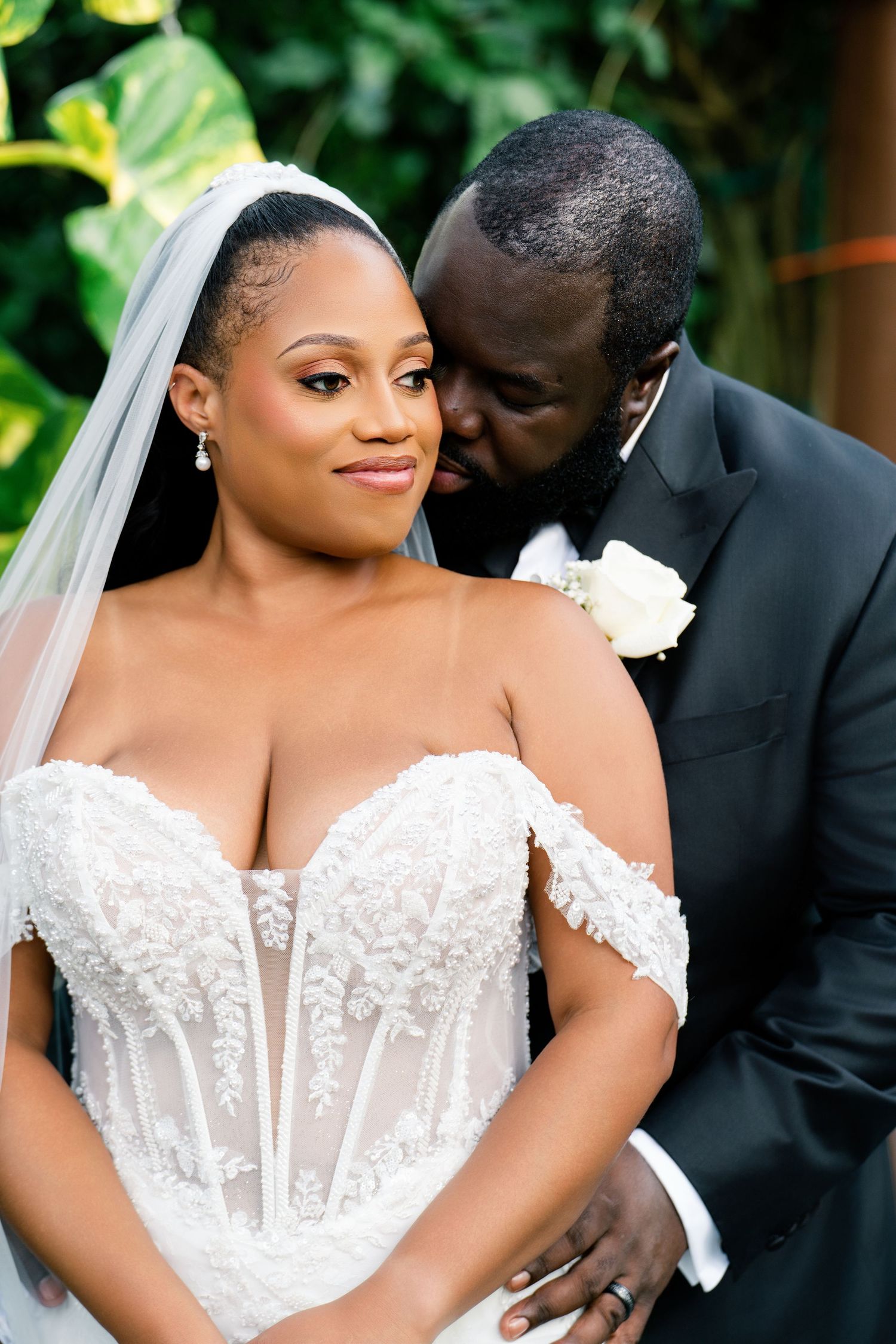 A bride in a lace off-shoulder gown with veil smiles as groom in tuxedo whispers in her ear outdoors.