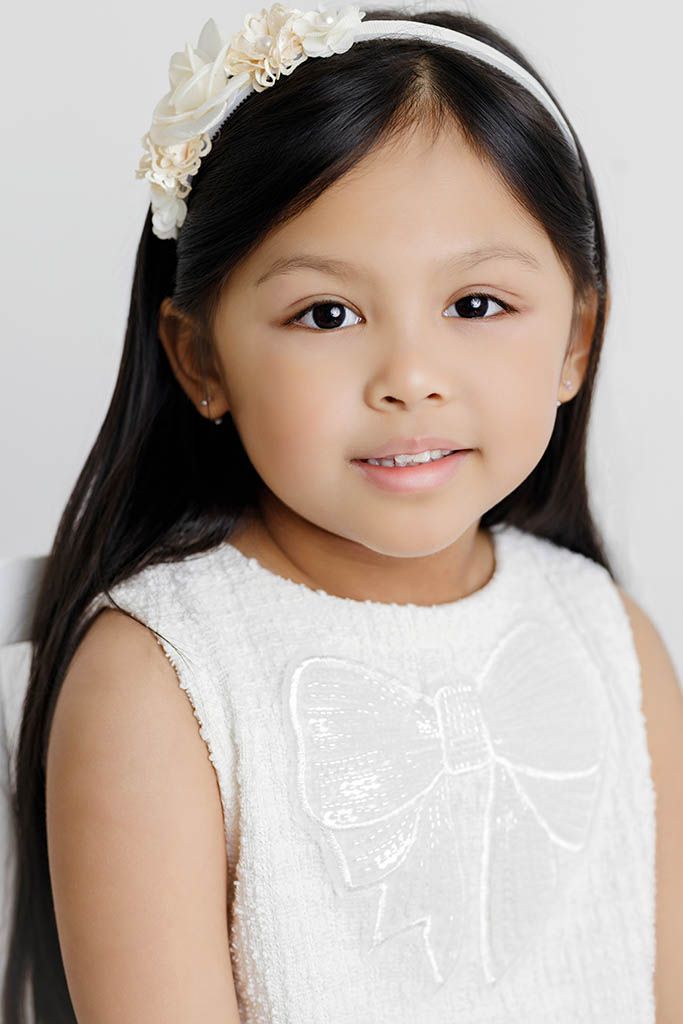Young girl in white textured dress with bow detail and floral headband, smiling for portrait photo.