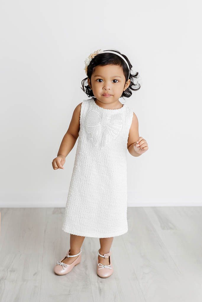 Toddler girl in white sleeveless dress with bow detail, floral headband, and pink Mary Jane shoes.