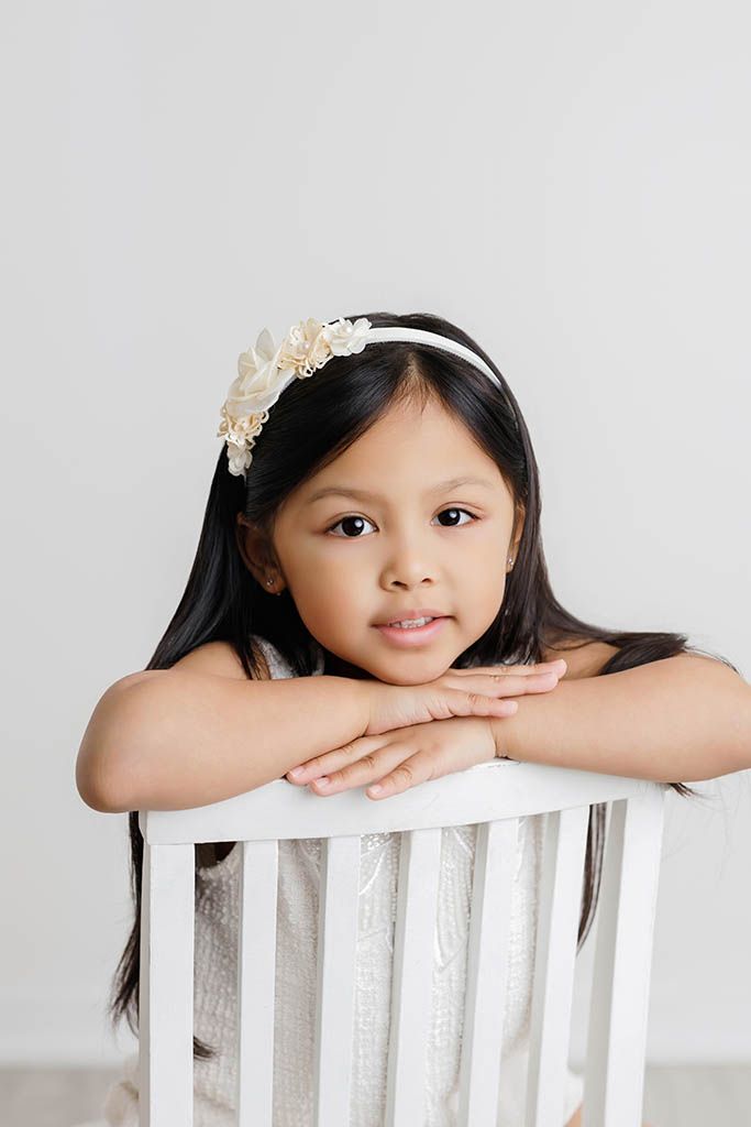 Young girl with long dark hair wearing a floral headband rests her chin on her hands over a white chair.