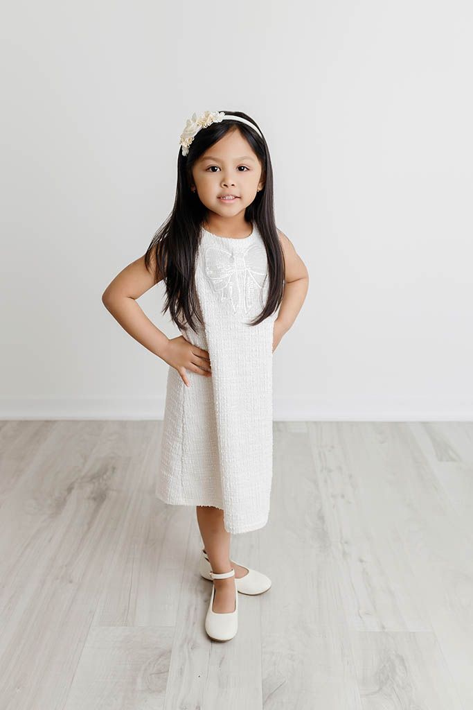 Young girl in white dress and floral headband poses confidently with hands on hips on light wood floor.