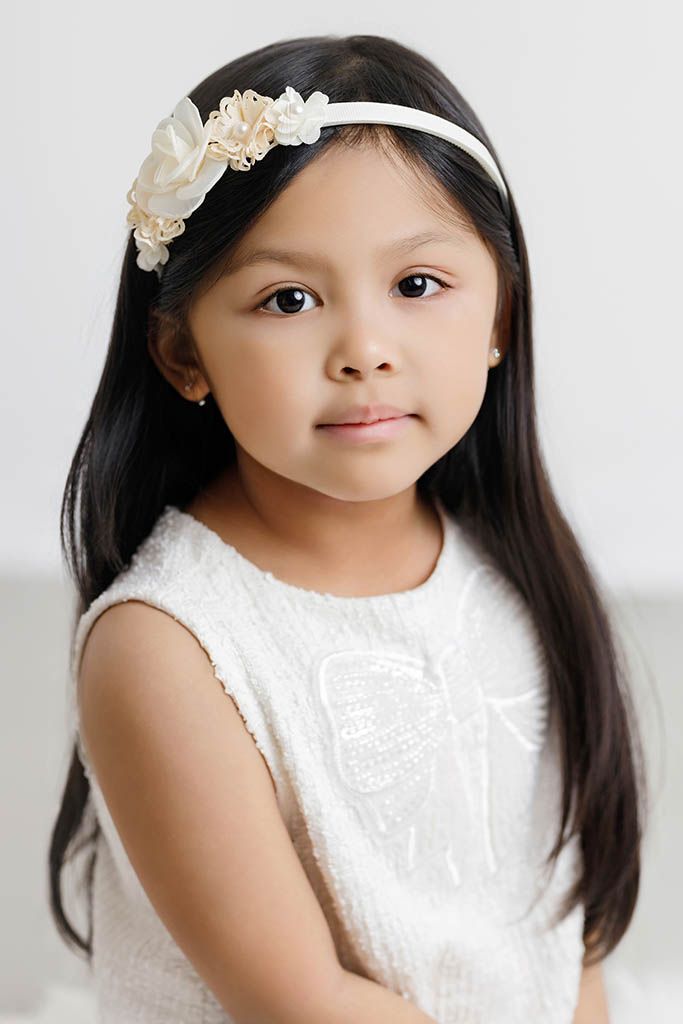 Young girl with long black hair wearing a white floral headband and white dress, posing for a portrait photo.
