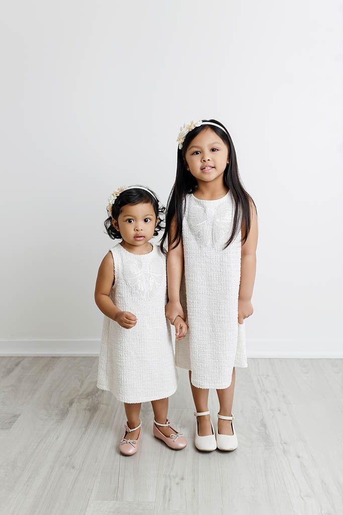 Two young girls in matching white dresses and floral headbands pose together for a portrait photo.