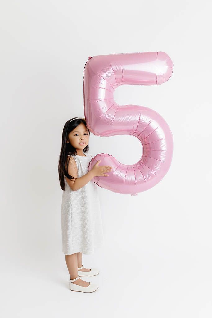 A young girl in a white dress holds a large pink number 5 balloon on a white background.