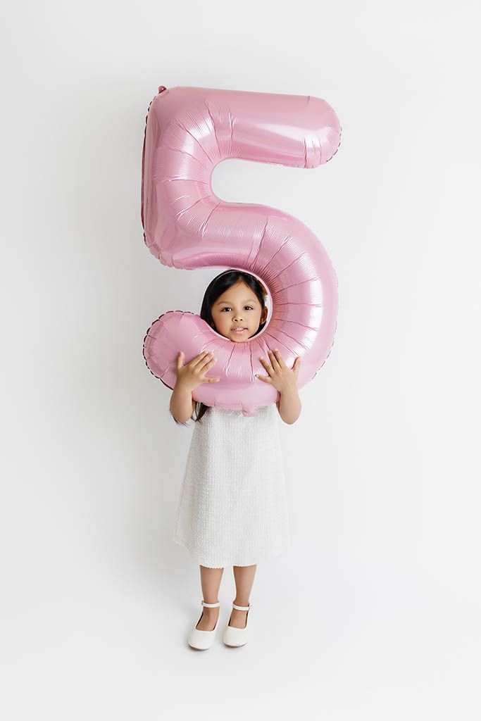 A young girl in a white dress holds a large pink number 5 balloon, celebrating her 5th birthday.